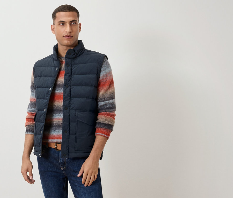 Un homme porte un gilet bleu marine sur un pull rayé, une ceinture marron et un jean.