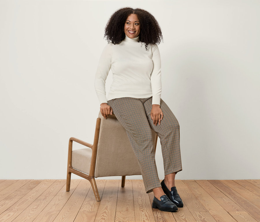 Une femme est assise sur un fauteuil beige, portant un pull à col roulé blanc, un pantalon pied-de-poule marron et des mocassins noirs.