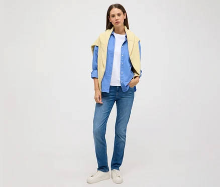 Une femme se tient sur un fond clair et porte un jean Angels «Cici Push-Up», coupe ajustée, une chemise bleue, un t-shirt blanc et un pull jaune.