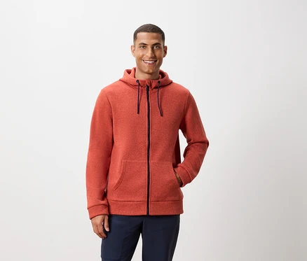 Un homme pose devant un fond blanc, portant un sweat à capuche zippé orange et un pantalon bleu marine.