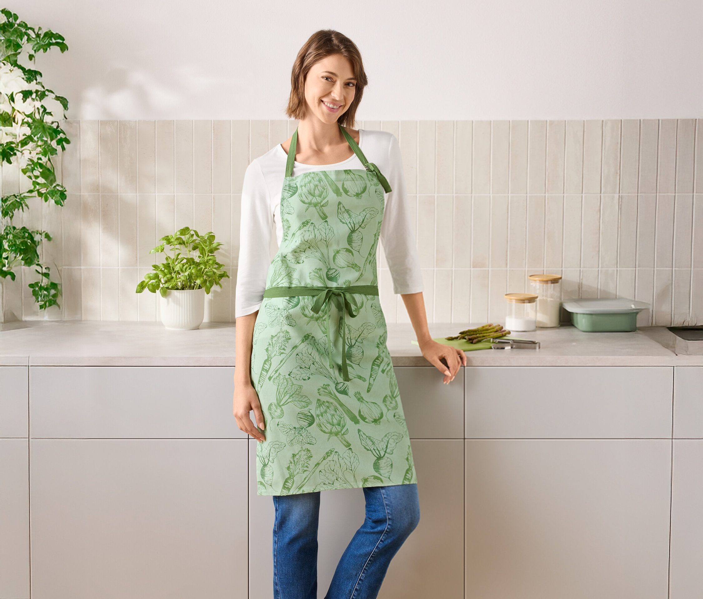 Une femme se tient dans une cuisine et porte un tablier vert à motifs de légumes.