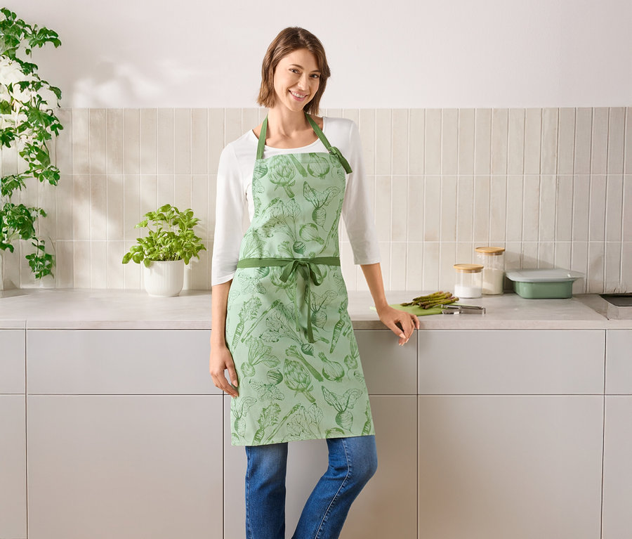 Une femme se tient dans une cuisine et porte un tablier vert à motifs de légumes.