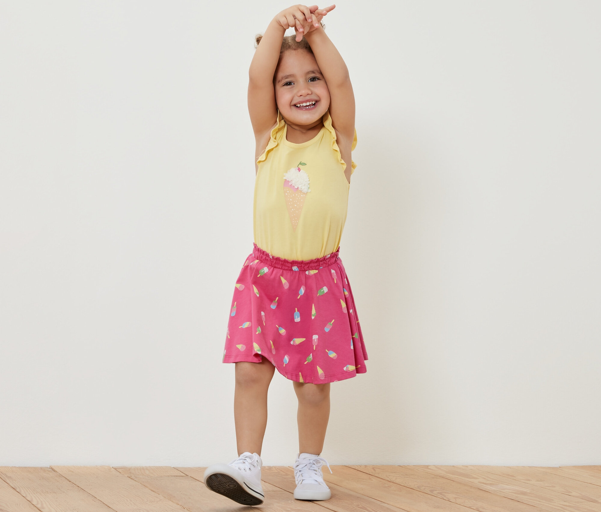Une fillette souriante se tient debout, les mains levées, portant un t-shirt jaune et une jupe en jersey pour enfants avec un pantalon à motif de glace.
