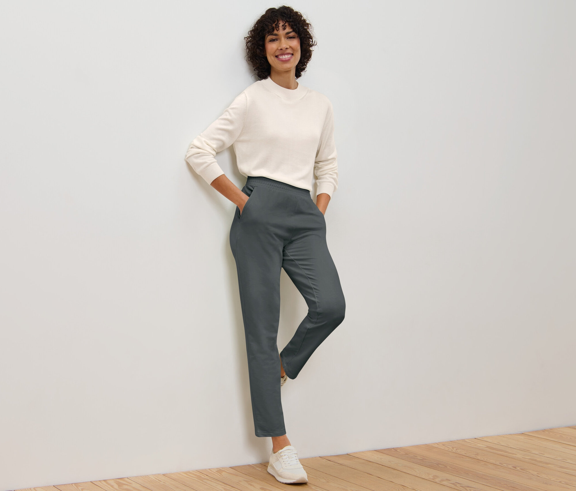 Une femme posant contre un mur, portant un pantalon de jogging gris et un pull.