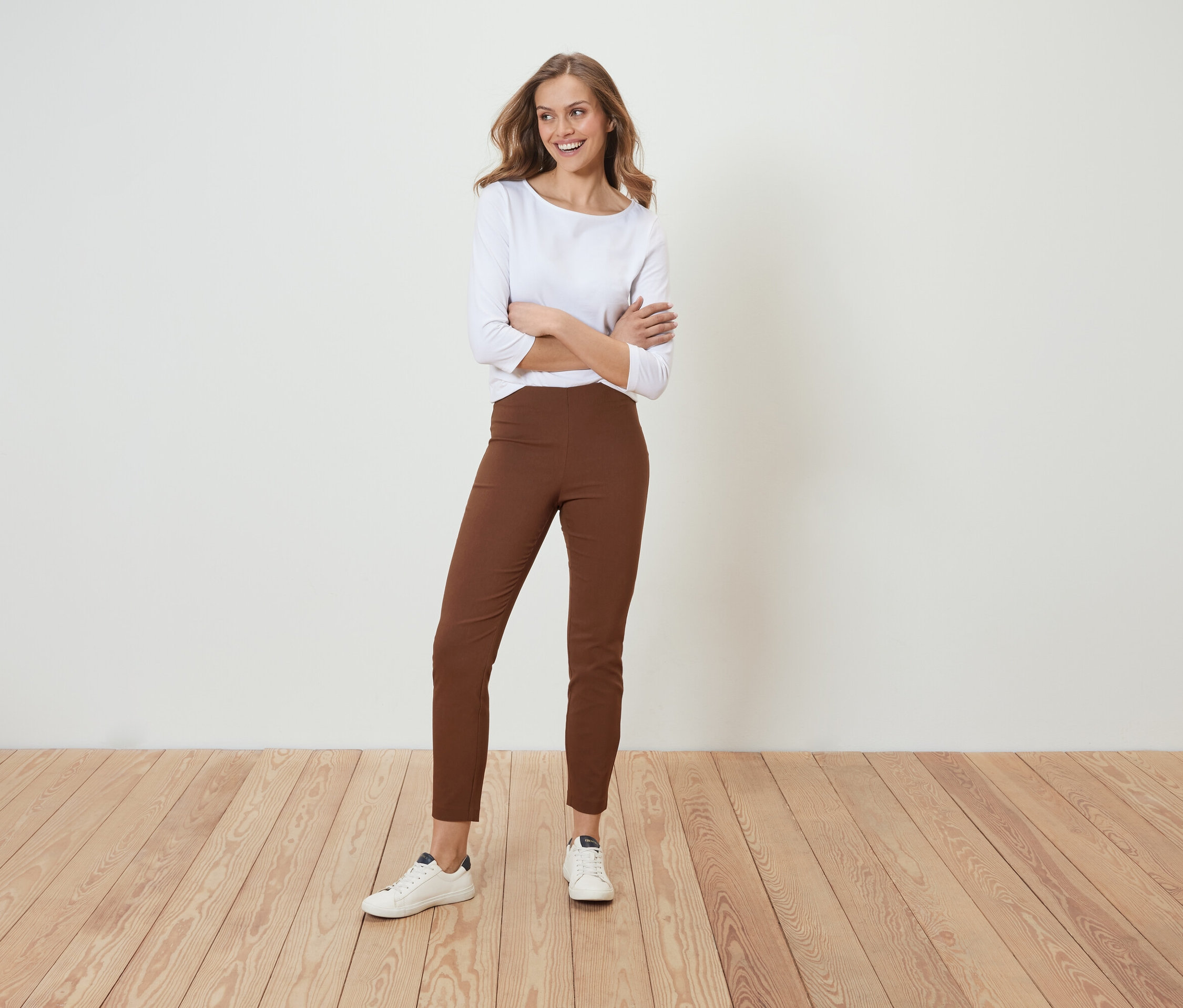 Une femme pose, les bras croisés, en pantalon marron et chemise blanche.