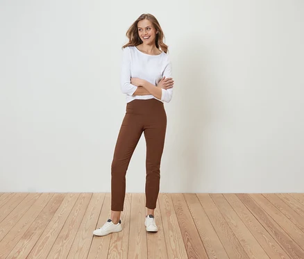 Une femme pose, les bras croisés, en pantalon marron et chemise blanche.