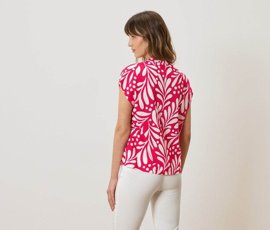 Femme vue de dos, portant un chemisier rose avec motif de feuilles blanches et un pantalon blanc.