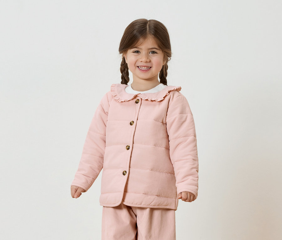 Fillette souriante avec des tresses portant une veste et un pantalon roses sur fond blanc.