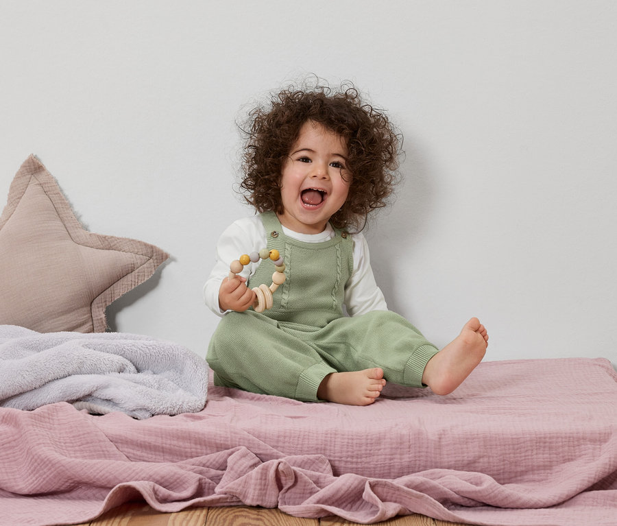 Un bébé souriant aux cheveux bouclés est assis sur une couverture rose et tient un jouet en bois.