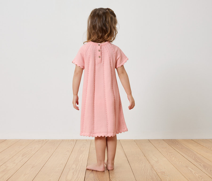 Une fillette est debout, pieds nus, dos à la caméra, portant une robe rose.