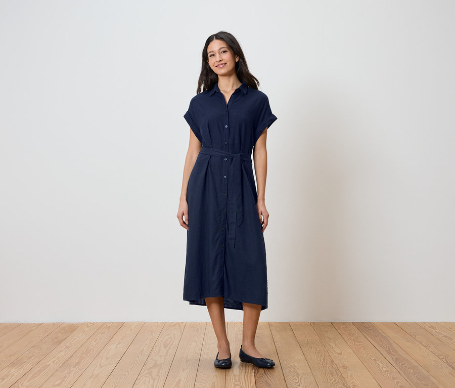 Une femme se tient devant un mur, portant une robe chemise bleu marine ceinturée et des ballerines.