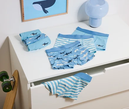 Sur une commode sont posés des boxers bleus pour enfants avec des motifs de requins et de rayures, à côté d'un skateboard et d'une lampe bleue.