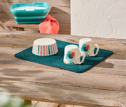 Sur une table en bois se trouve un égouttoir turquoise sur lequel reposent un bol à rayures roses et turquoises et deux tasses peintes. À l'arrière-plan, on aperçoit un bol turquoise pliable rempli et un tissu orange.