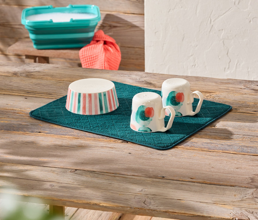 Sur une table en bois se trouve un égouttoir turquoise sur lequel reposent un bol à rayures roses et turquoises et deux tasses peintes. À l'arrière-plan, on aperçoit un bol turquoise pliable rempli et un tissu orange.