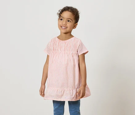 Une jeune fille souriante porte une chemise rose et blanche à rayures et un jean bleu.