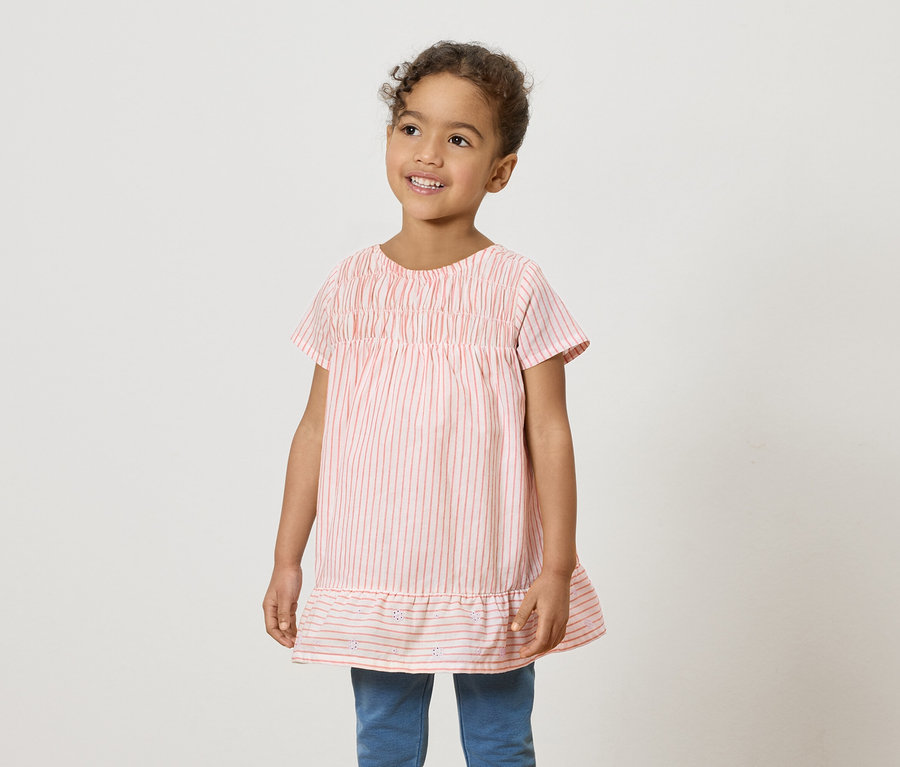 Une jeune fille souriante porte une chemise rose et blanche à rayures et un jean bleu.