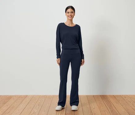 Une femme aux cheveux foncés portant un haut à manches longues bleu marine et un pantalon assorti devant un mur blanc.