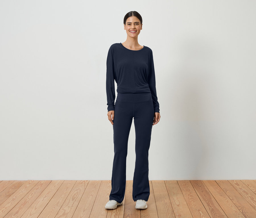 Une femme aux cheveux foncés portant un haut à manches longues bleu marine et un pantalon assorti devant un mur blanc.