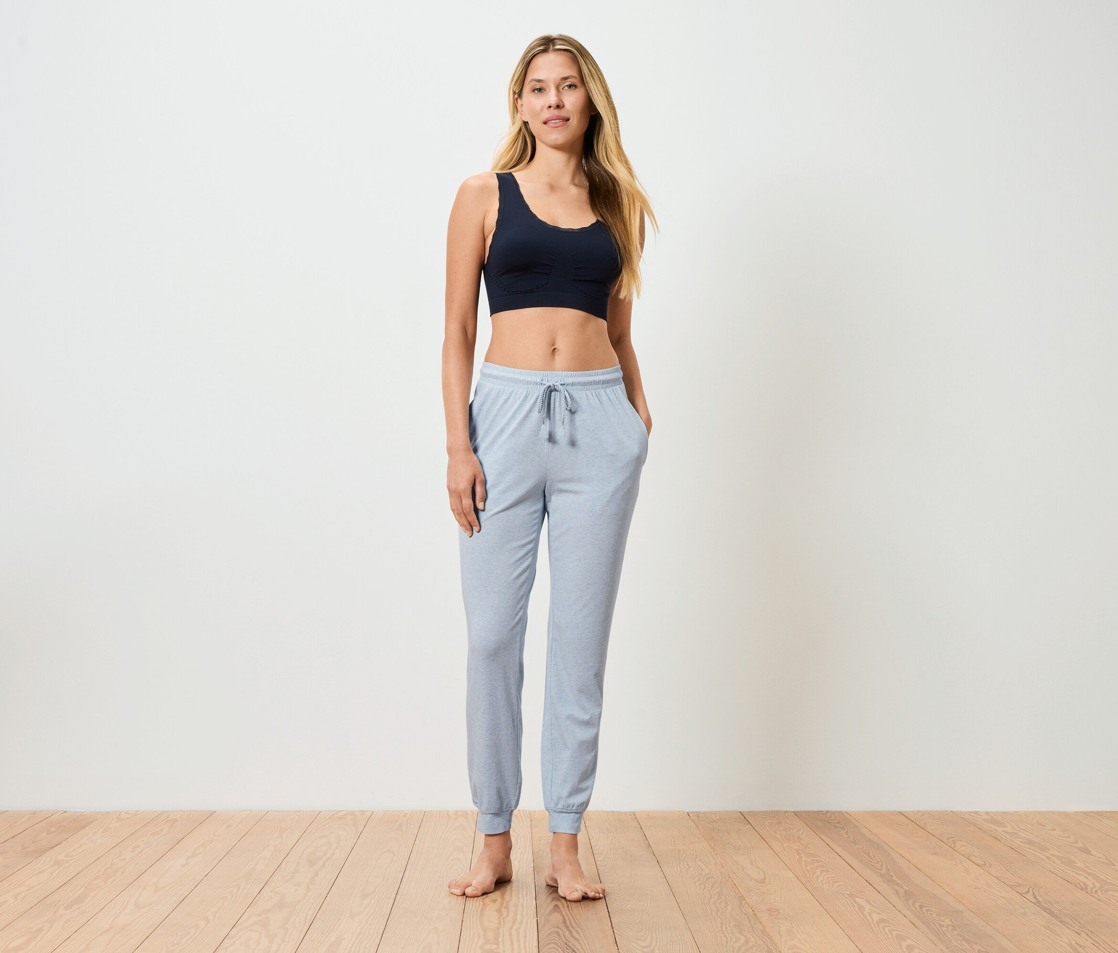 Femme debout pieds nus avec un pantalon de jogging bleu clair et un soutien-gorge de sport foncé devant un mur blanc.