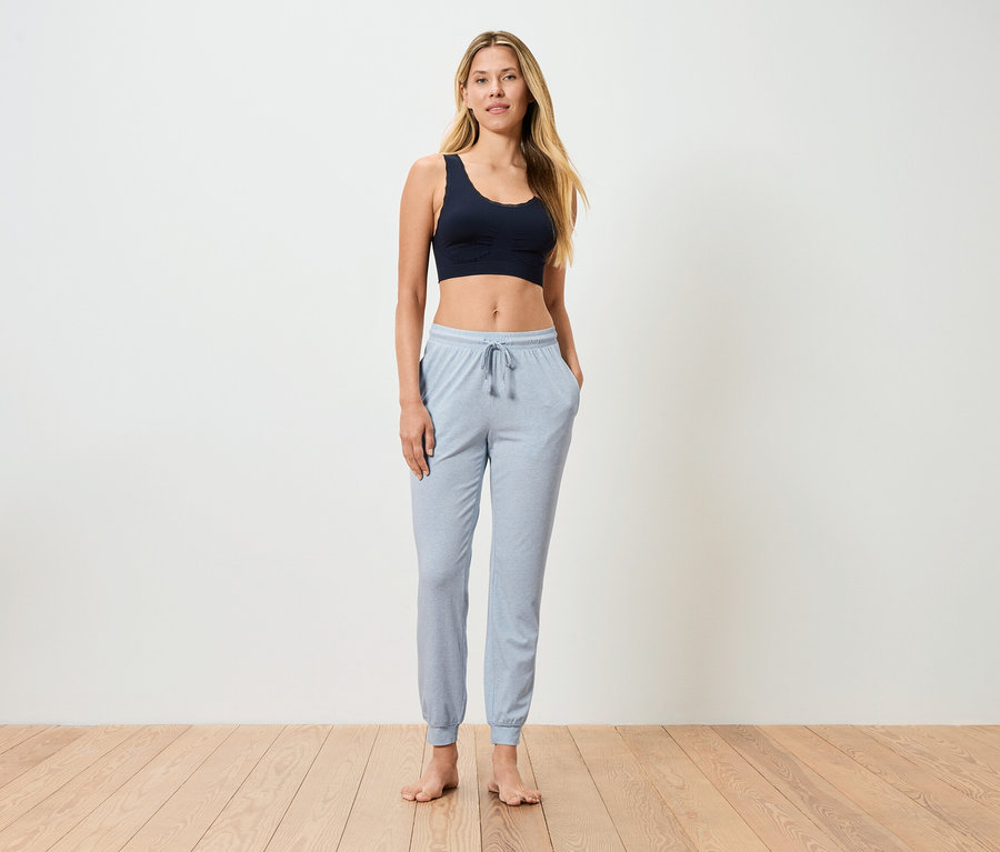 Femme debout pieds nus avec un pantalon de jogging bleu clair et un soutien-gorge de sport foncé devant un mur blanc.