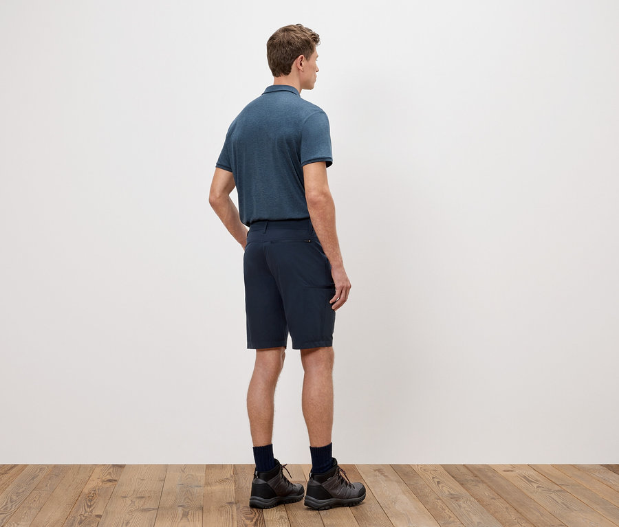 Un homme, vu de dos, porte un polo bleu, un short et des chaussures de randonnée devant un mur blanc.