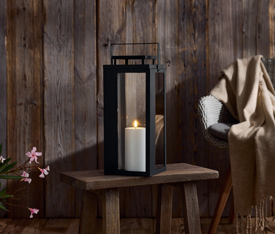 Une lanterne noire avec une bougie blanche sur une petite table en bois. En arrière-plan, un mur en bois et une chaise avec une couverture.