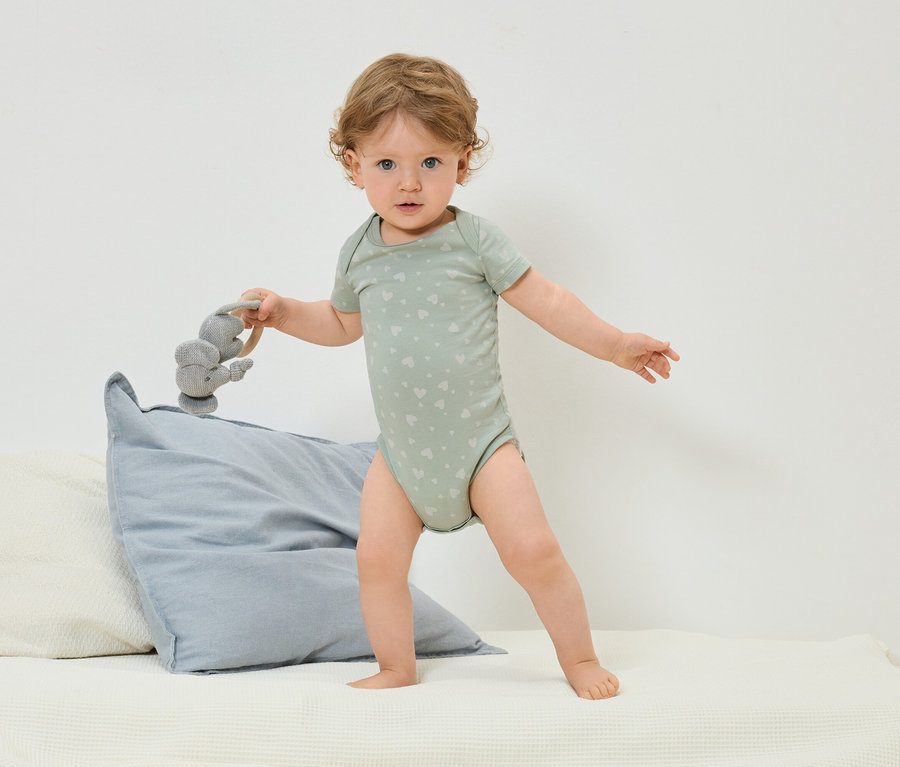 Un bébé portant un body vert à motif de cœurs se tient debout sur un lit et tient un jouet éléphant gris.
