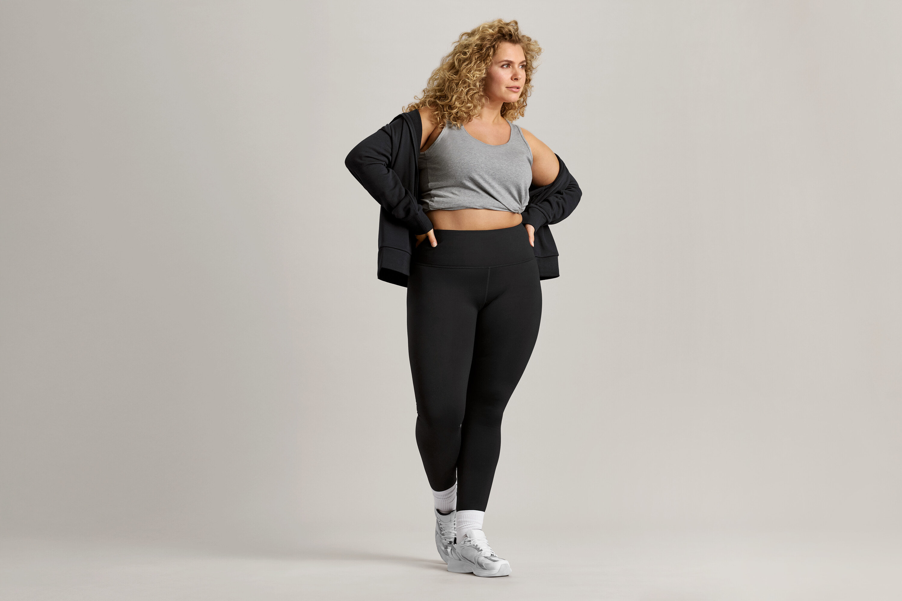 Une femme aux cheveux bouclés porte un débardeur gris, une veste noire et un legging noir.