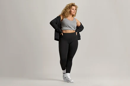 Une femme aux cheveux bouclés porte un débardeur gris, une veste noire et un legging noir.