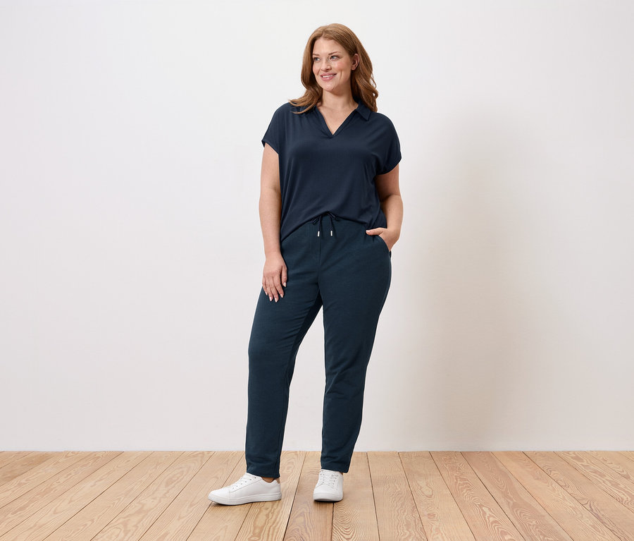 Une femme se tient devant un mur, elle porte un t-shirt bleu marine, un pantalon bleu marine et des baskets blanches.