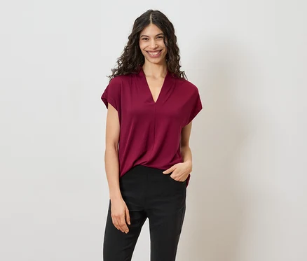 Une femme aux cheveux bouclés et foncés porte un t-shirt bordeaux et un pantalon noir sur un fond blanc.