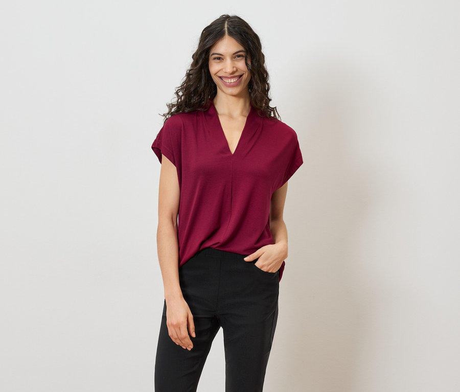 Une femme aux cheveux bouclés et foncés porte un t-shirt bordeaux et un pantalon noir sur un fond blanc.