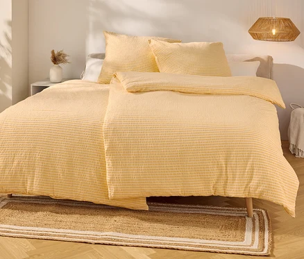 Des draps jaunes rayés recouvrent un lit avec deux oreillers. Un tapis est posé devant.