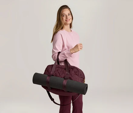 Une femme portant un pull rose et un pantalon bordeaux porte un sac de sport bordeaux avec un tapis de yoga noir sur l'épaule.