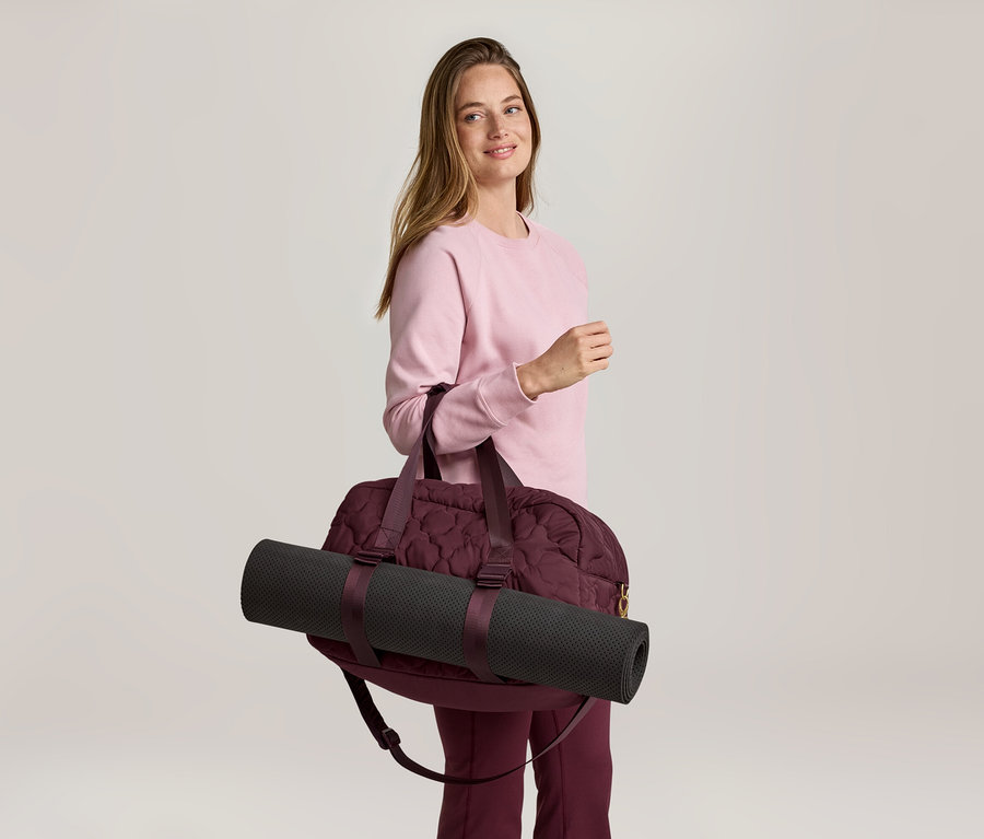 Une femme portant un pull rose et un pantalon bordeaux porte un sac de sport bordeaux avec un tapis de yoga noir sur l'épaule.