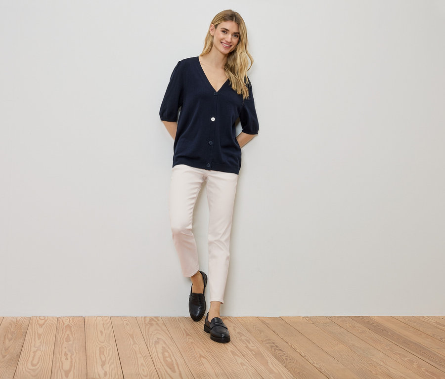 Une femme se tient contre un mur, portant un cardigan bleu marine, un pantalon clair et des chaussures noires.