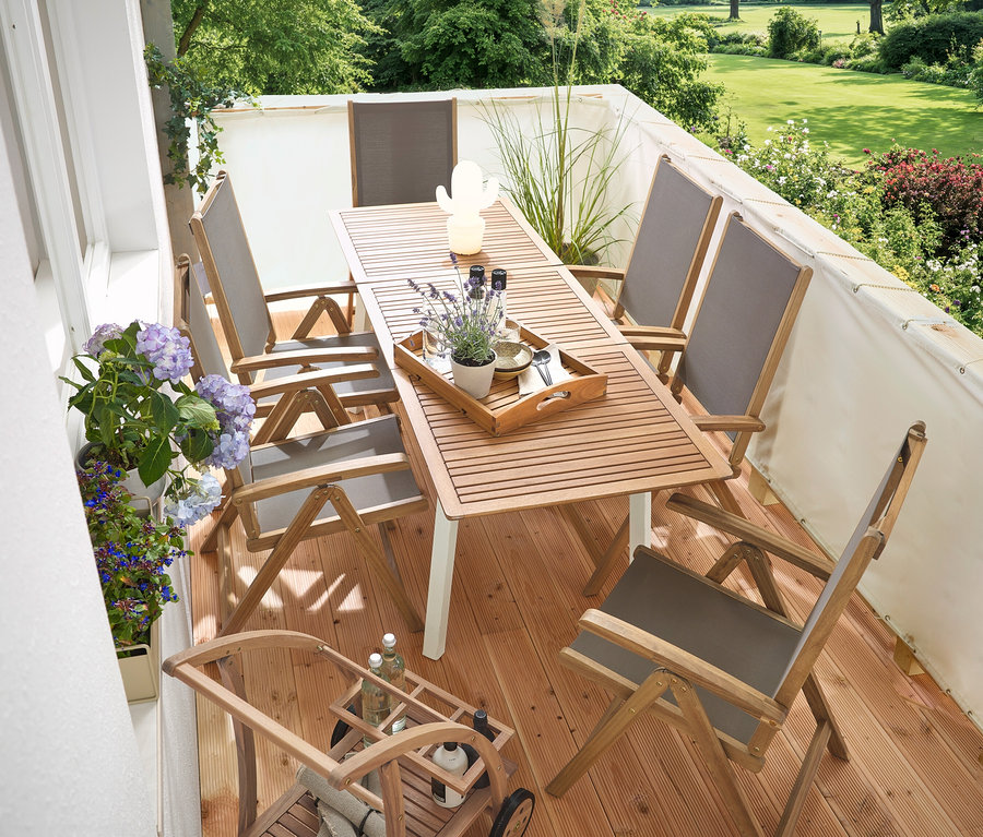Sur un balcon se trouvent une table de jardin étroite extensible »Liska«, un brise-vue et brise-vent de balcon crème, un barwagen mit abnehmbarem Tablett et une lampe de table à LED d'extérieur rechargeable en forme de cactus.
