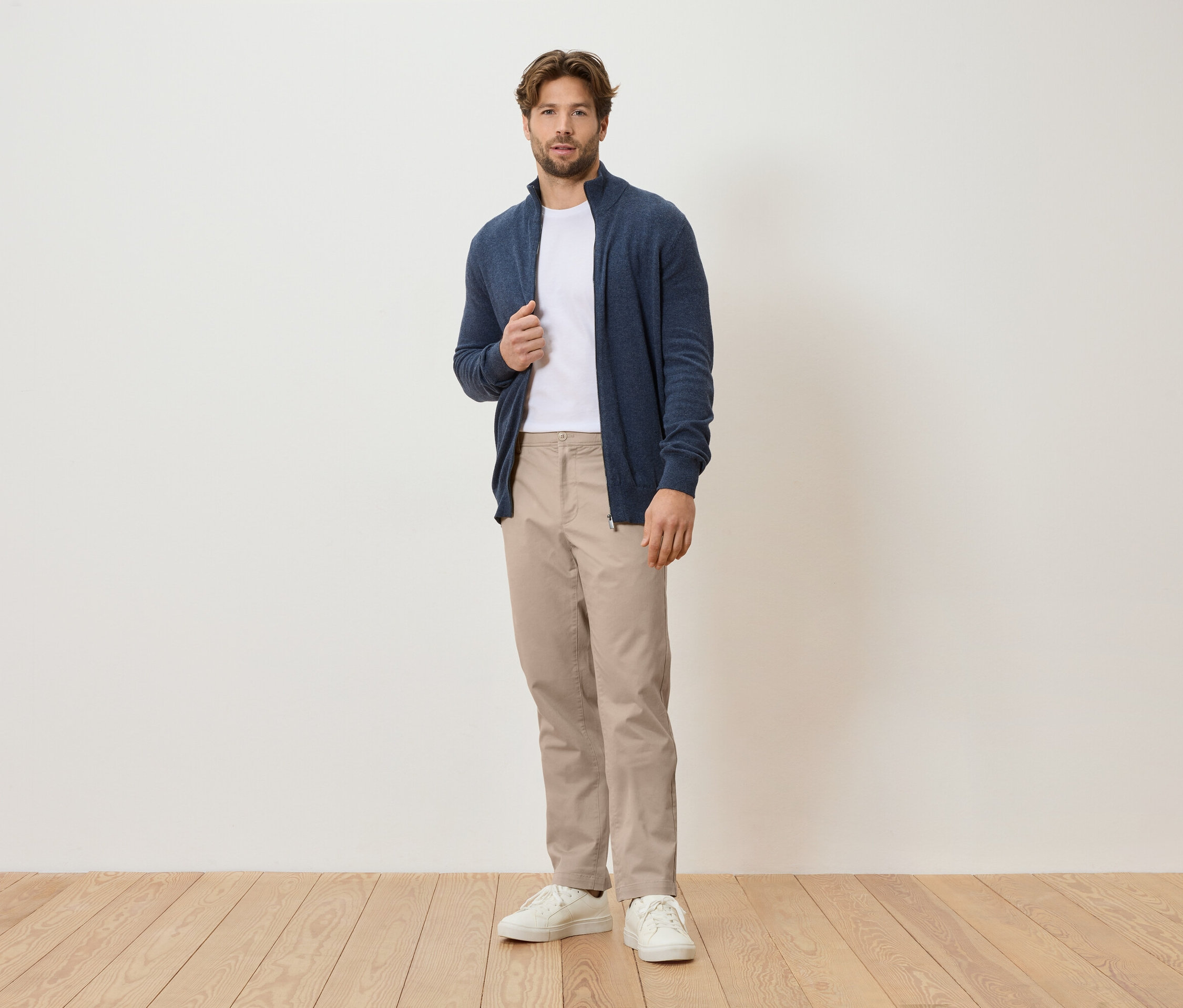 Un homme se tient devant un mur blanc, il porte un cardigan bleu marine, un t-shirt blanc, un pantalon beige et des baskets blanches.