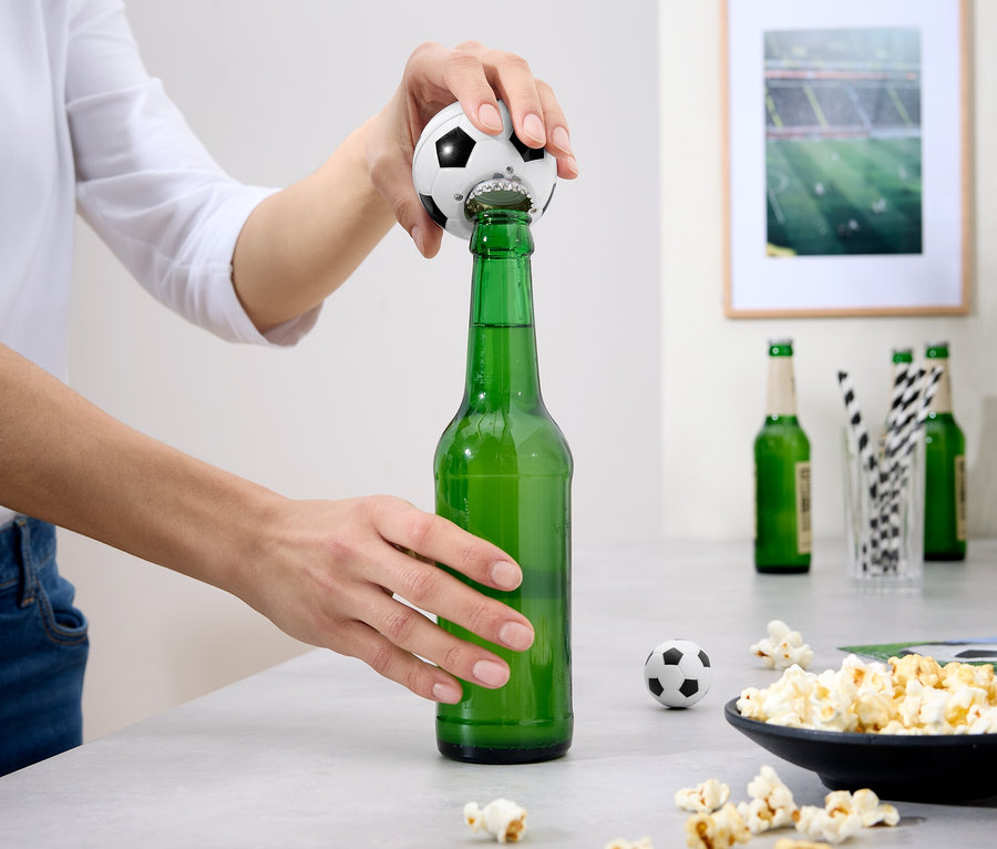 Une personne ouvre une bouteille de bière verte avec un ouvre-bouteille en forme de ballon de football. Il y a du pop-corn et d'autres bouteilles sur la table.
