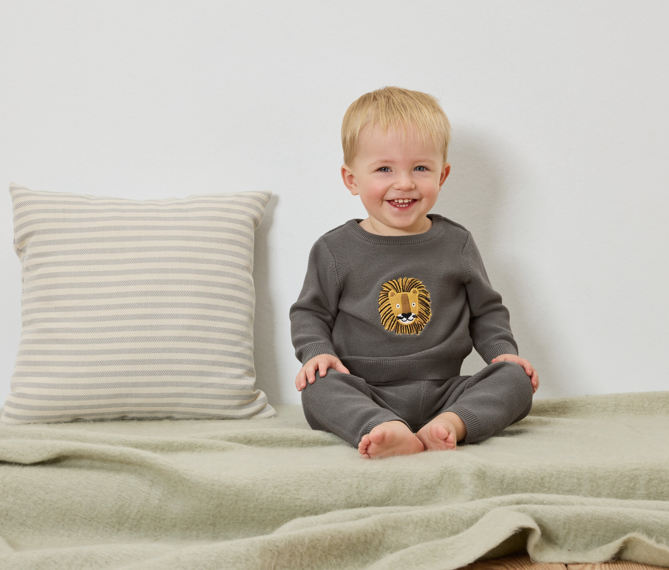 Un jeune enfant blond souriant est assis sur une couverture vert clair à côté d'un coussin rayé.