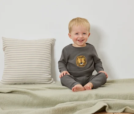 Un jeune enfant blond souriant est assis sur une couverture vert clair à côté d'un coussin rayé.