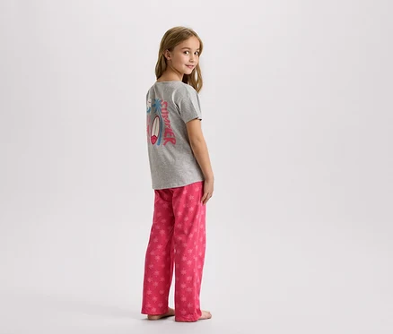 Une jeune fille se tient debout, portant un t-shirt gris avec un motif estival et un pantalon rose à palmiers.