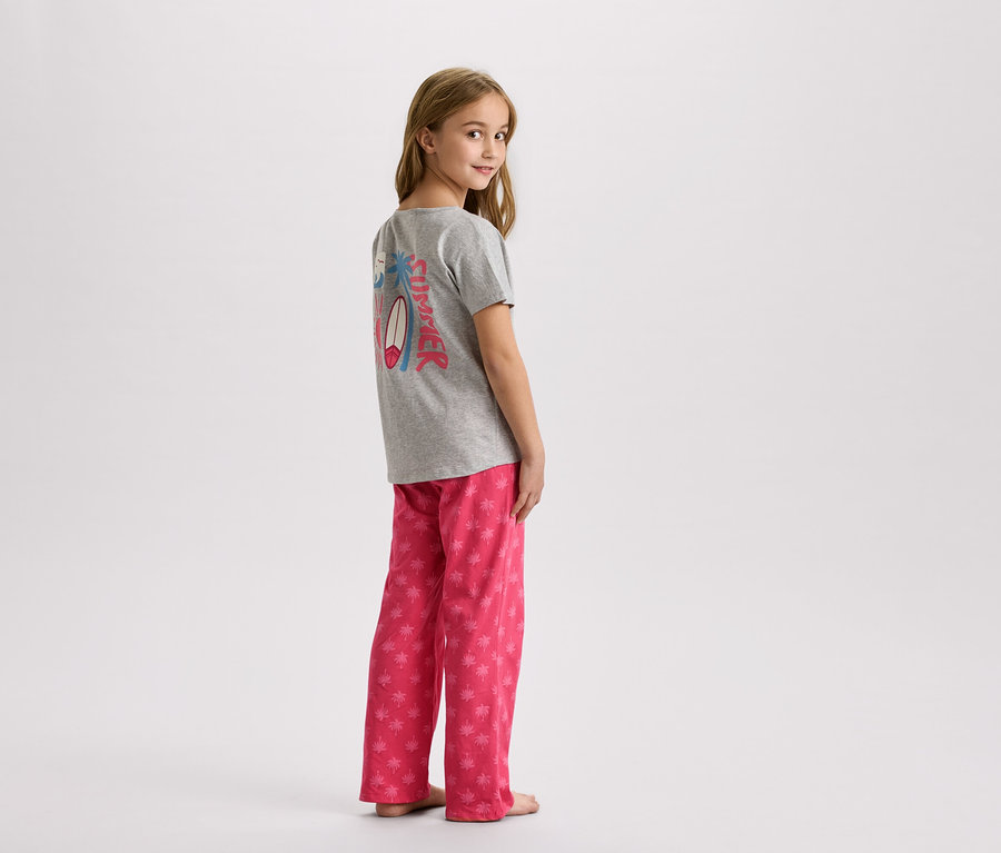 Une jeune fille se tient debout, portant un t-shirt gris avec un motif estival et un pantalon rose à palmiers.