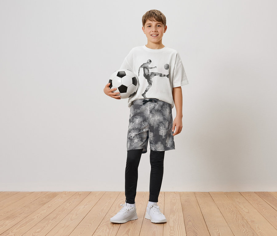 Un garçon tient un ballon de football dans sa main gauche. Il porte un t-shirt avec un joueur de football, un short et des leggings, ainsi que des baskets blanches.
