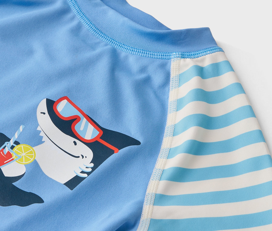Gros plan d'un haut de bain bleu clair pour enfant avec un requin portant des lunettes de soleil et tenant un cocktail, et des manches rayées.