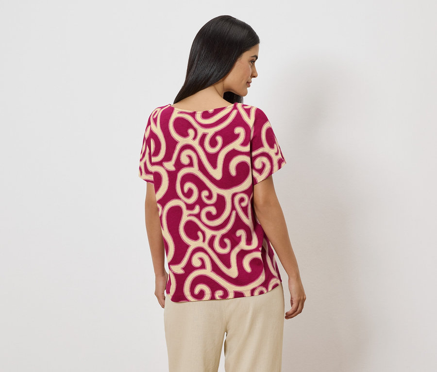 Femme vue de dos, portant un t-shirt bordeaux avec des motifs beiges et un pantalon beige sur fond blanc.