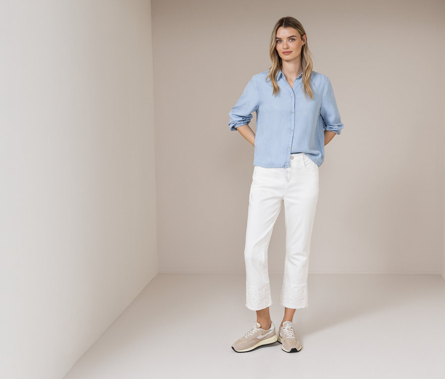 Une femme se tient dans une pièce et porte une chemise bleu clair, un jean blanc avec de la dentelle et des baskets beiges.