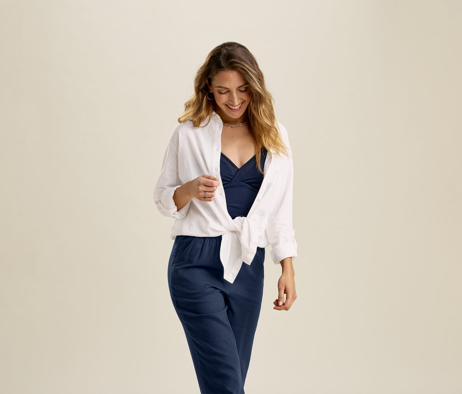 Une femme porte une blouse longue en lin blanche nouée sur le devant, un haut bleu et un pantalon bleu.