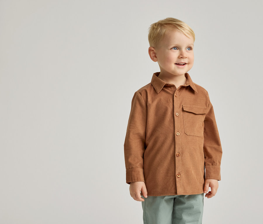 Un garçon souriant aux cheveux blonds, vêtu d'une chemise en velours côtelé marron et d'un pantalon vert, se tient devant un fond gris clair.