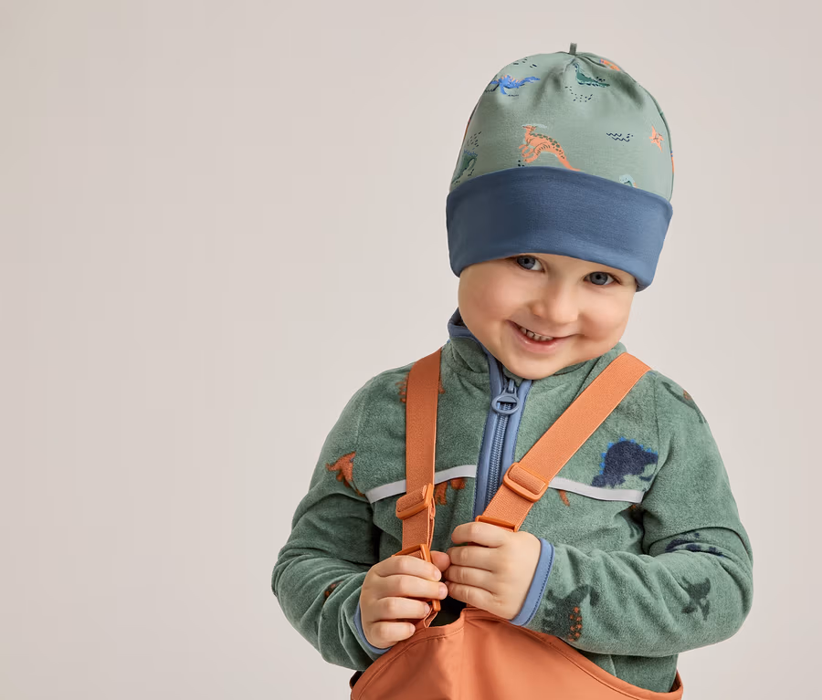 Un garçon souriant portant un bonnet à motifs de dinosaures, un pull en polaire vert et une salopette orange pose devant un fond clair.